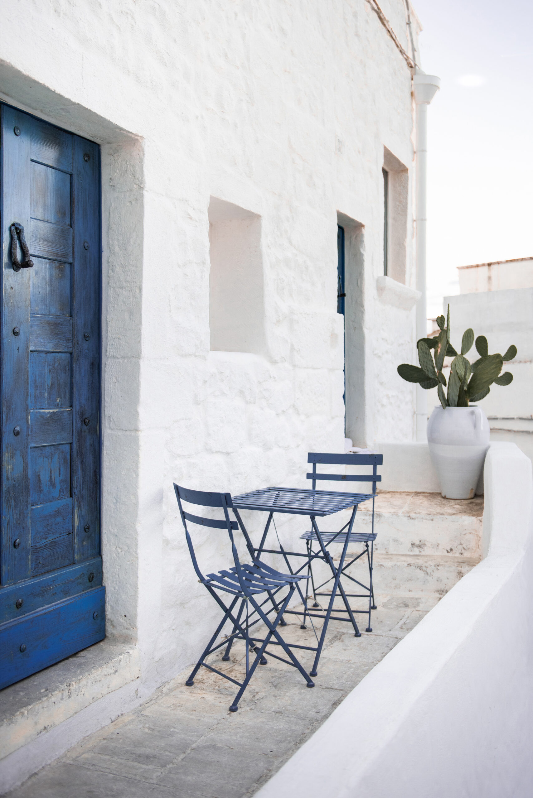 white building in Greece with a dark blue door, showing the front porch with a dark blue metal table and matching metal chairs with a cactus in a planter in the background