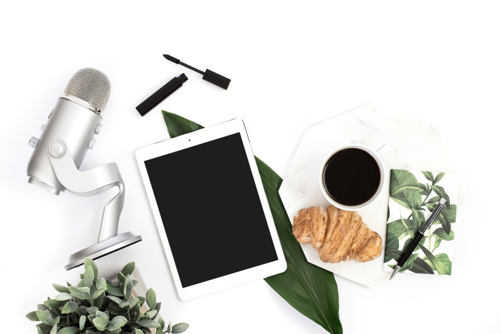Flat lay of a creative workspace featuring a silver microphone, a blank-screen tablet, an open mascara tube, a croissant, a cup of black coffee, and a leafy notebook with a pen. The setup is styled with greenery on a white background, evoking a cozy, productive morning vibe.